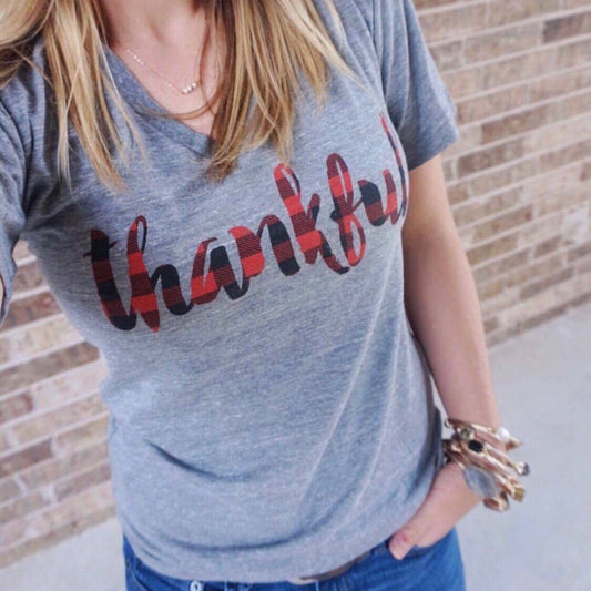 A person with light brown hair, necklace, and bracelets stands before a brick wall wearing the Thankful Plaid • Tee, a gray unisex poly/cotton blend shirt featuring red and black Thankful plaid lettering.