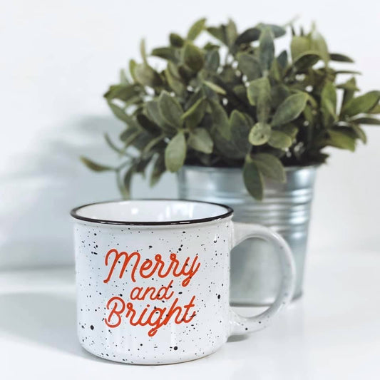 The Merry & Bright • Campfire Ceramic Mug features a white speckled ceramic design with red lettering, displayed in front of a potted green plant in a metal container on a white surface. Hand-wash only.