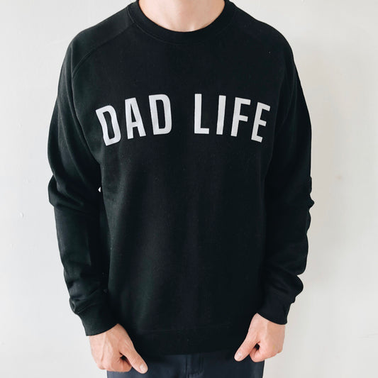 A person wears the Dad Life Block Pullover, a unisex black fleece-lined sweatshirt featuring bold white DAD LIFE text on the front, shown against a plain light-colored wall.