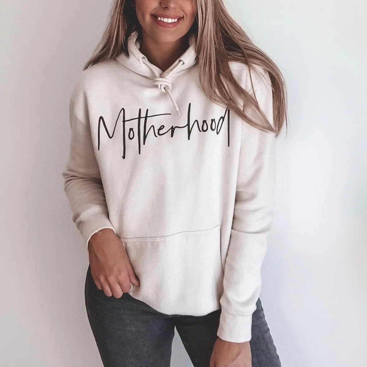 A woman wears the Motherhood • Sand Hoodie, featuring Motherhood in bold black script on the front. With its unisex fit, this light-colored sweatshirt offers comfort and versatility. She smiles softly against a plain white background.