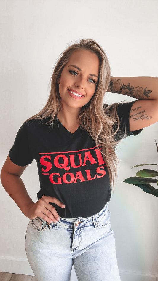 A smiling woman with long blonde hair stands indoors, wearing light jeans and the comfy Squad Goals • Youth tee. One hand is on her hip, the other behind her head, showing a visible forearm tattoo.