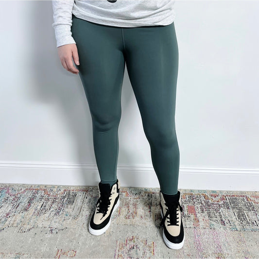 A person wearing Jade Leggings, a light gray long-sleeve shirt, and high-top beige and black sneakers stands on a multicolored rug against a white wall.