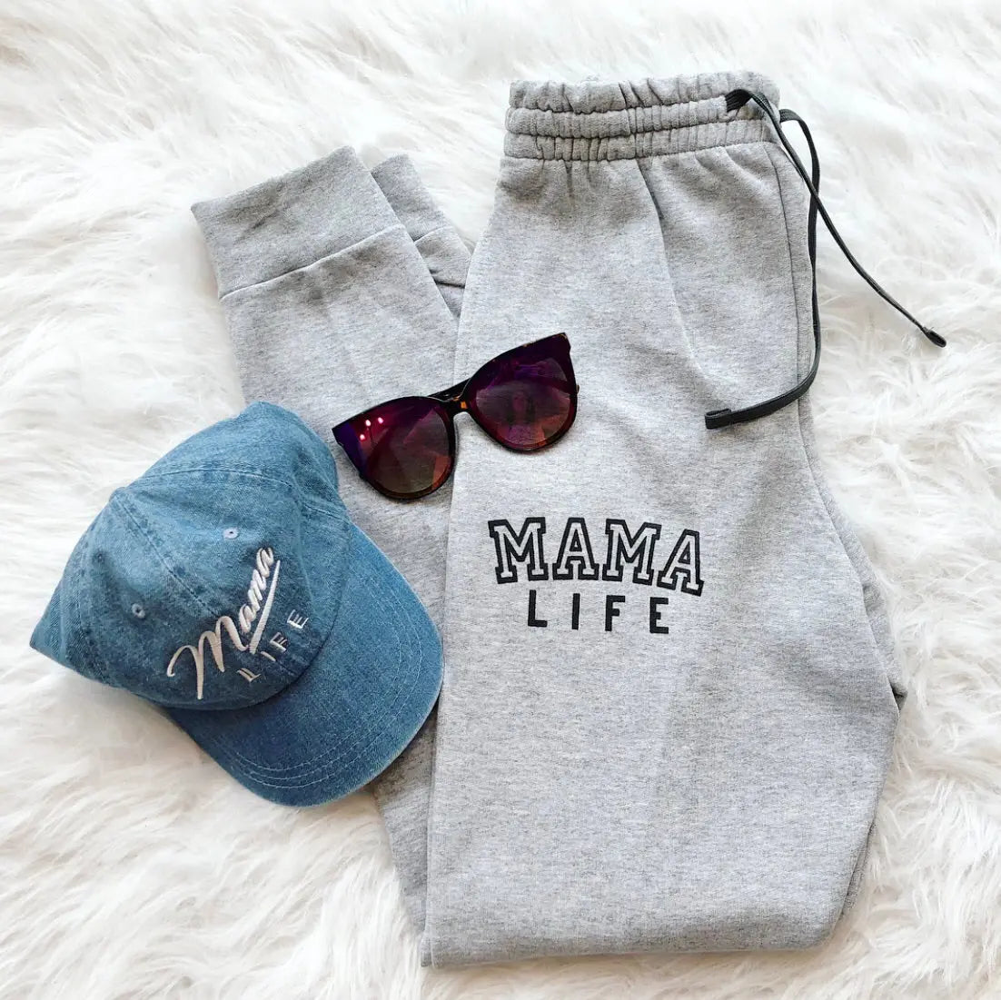 The Mama Life Joggers in gray, styled with dark sunglasses and a blue Mama Life embroidered cap, are displayed on a white, fluffy surface for the ultimate cozy loungewear vibe.