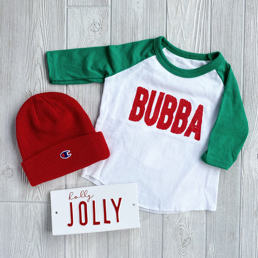 A Bubba Holiday Raglan with green sleeves and red “BUBBA” lettering, paired with a red Champion beanie and a white “holly JOLLY” sign, displayed on a light wood surface.