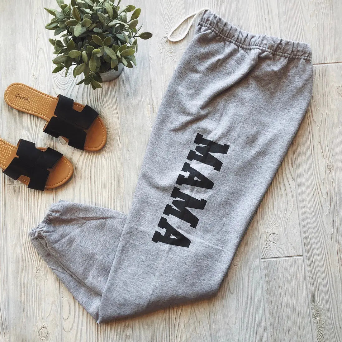 The MAMA Sweats • gray, featuring bold black lettering, are styled on a light wood floor beside black sandals and a small potted plant—ideal for cozy loungewear.