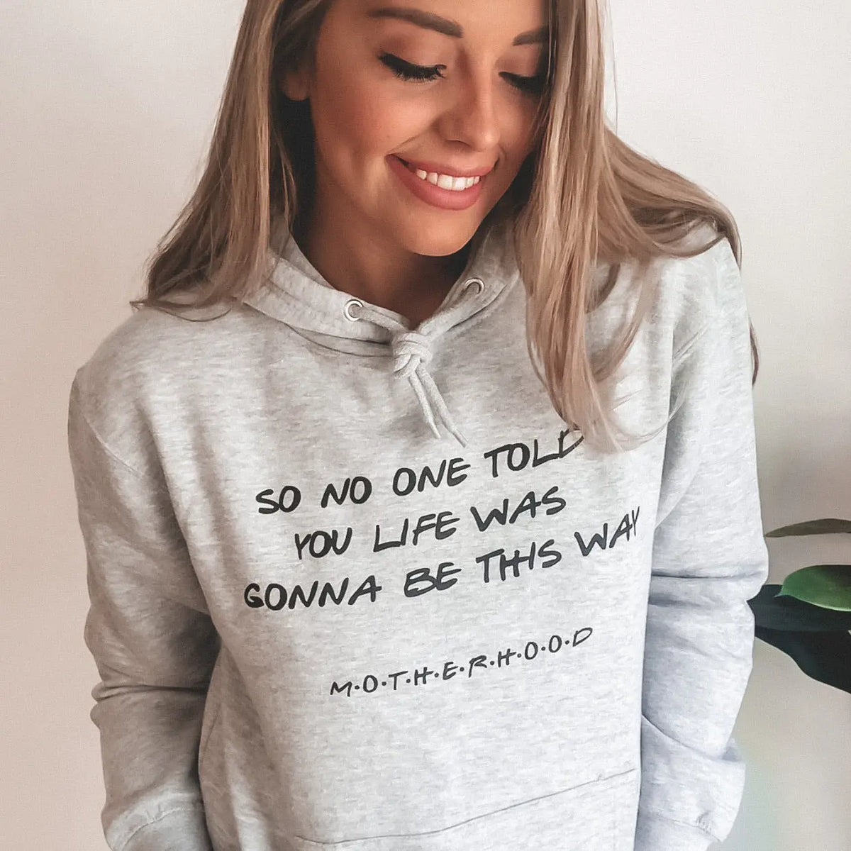 A smiling woman wears the No One Told You • Gray Hoodie, featuring a Friends parody and the text SO NO ONE TOLD YOU LIFE WAS GONNA BE THIS WAY MOTHERHOOD on the front.