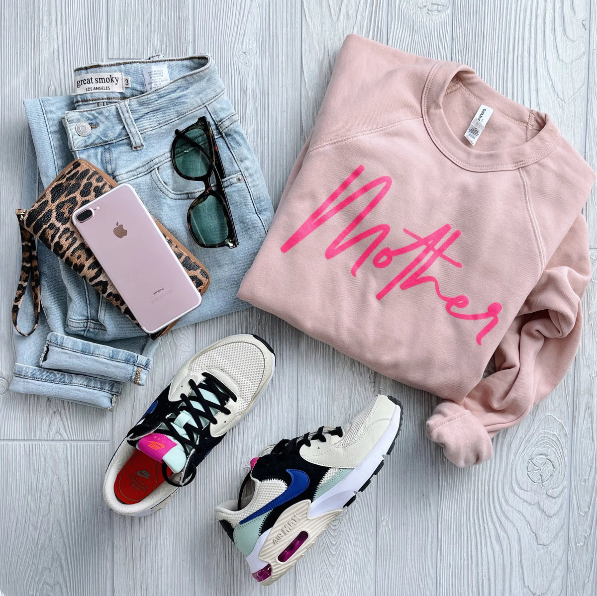 Flat lay of a casual outfit: light blue jeans, Mother • Peach Pullover sweatshirt, leopard print wallet, pink smartphone, sunglasses, and white, black, and pink sneakers on a light wooden floor.
