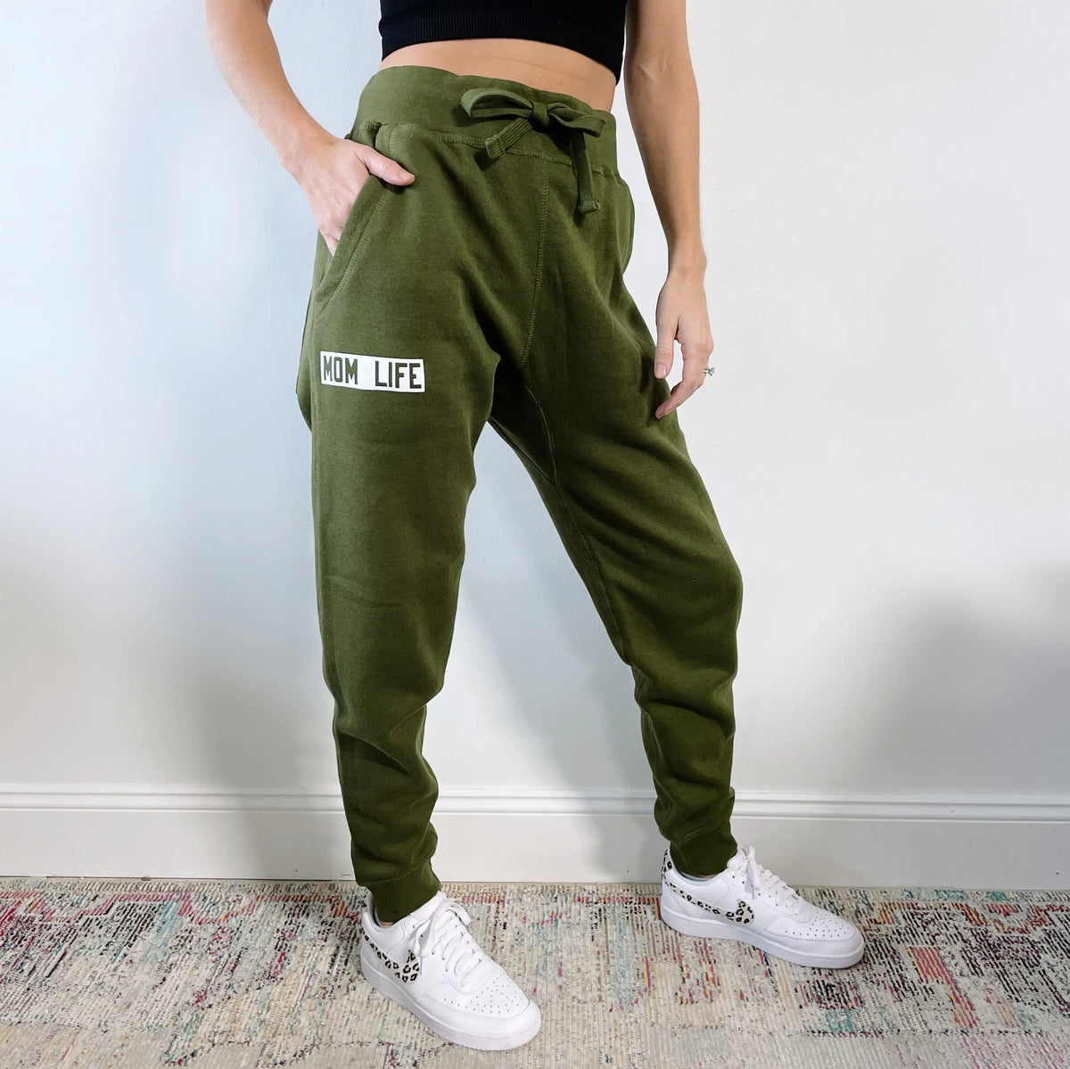 Someone wearing the Mom Life • Olive Joggers with MOM LIFE on the left thigh, a black crop top, and white sneakers stands on a patterned rug against a white wall.