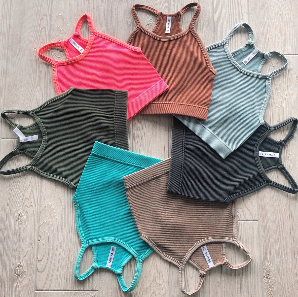 Six Luna Vintage Crop tops from the Aurora Collection—pink, brown, green, teal, gray, and charcoal—are arranged in a circle on light wood. Each ribbed, high-neck sleeveless top is made of stretchy fabric and pairs with matching shorts seen partially.