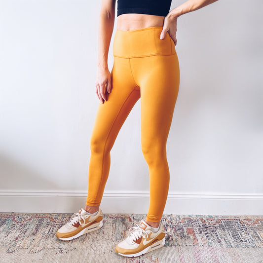 Person stands indoors against a white wall, wearing a black crop top, Lennon Leggings in yellow, and white and orange Nike sneakers. Face is not visible.
