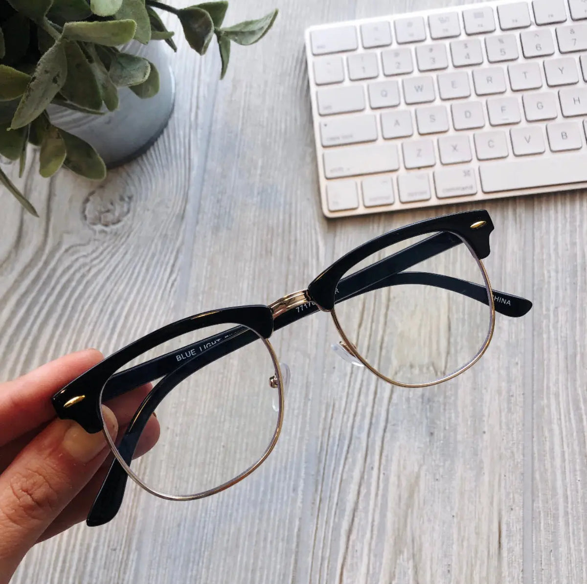 A hand holds Parker Blue Light Glasses—sleek black-rimmed eyewear that reduces blue light and eye strain—above a light wood desk, with part of a white keyboard and a potted plant in the background. More colors available!.