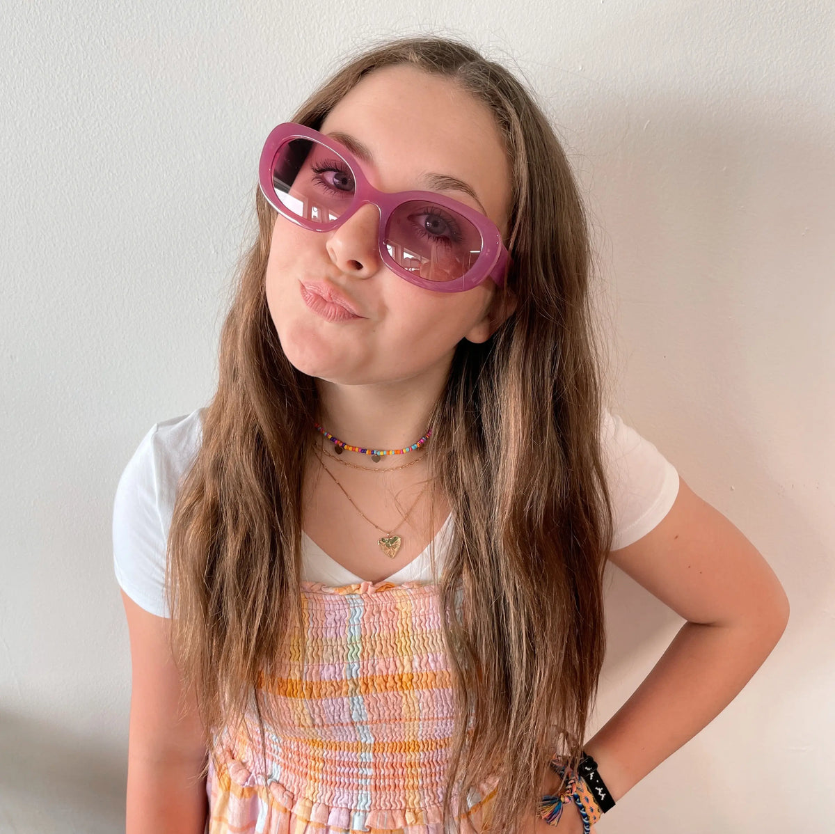 A girl with long brown hair wears Poppy Sunglasses • Adult • More Colors! in pink, paired with a white tee and pastel dress. She pouts playfully with one hand on her hip, standing against a plain white wall.