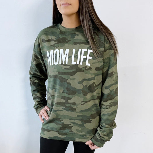 A woman wears the Mom Life Block • Discontinued Camo Pullover, a green camo sweatshirt with MOM LIFE in white letters and a comfy unisex fit by Independent, standing against a plain white background.