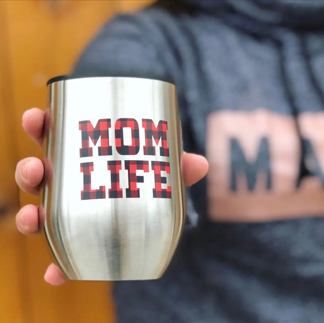 Someone in a gray MAM sweatshirt holds the Mom Plaid • Wine Tumbler, which features a bold red and black buffalo plaid Mom Life print. The blurred background highlights the eye-catching tumbler and the hand holding it.
