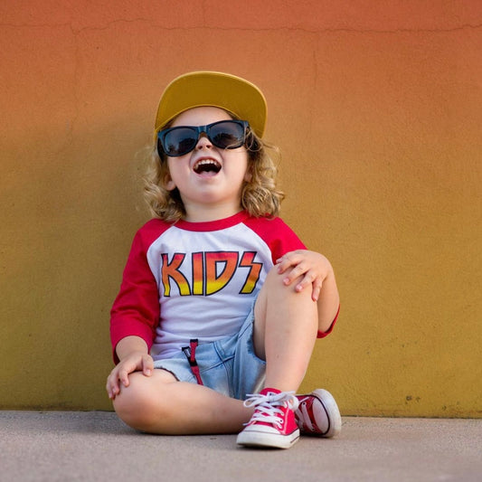 A young child with curly hair smiles while wearing a yellow cap, black sunglasses, denim shorts, red sneakers, and a true-to-size KIDS • Raglan shirt against an orange-yellow wall, showing off a cool little rocker style.