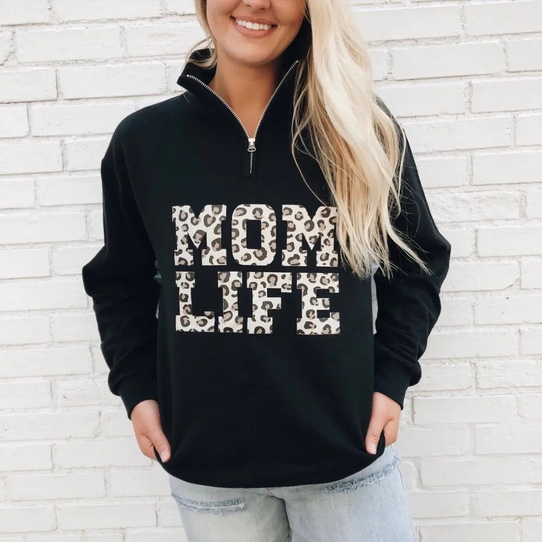 A woman with long blonde hair models the Mom Life Cheetah • Quarter Zip, featuring bold cheetah print MOM LIFE lettering on the front and a 1/4 zip design, standing against a white brick wall with her hands in her pockets.