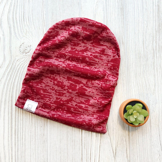A Red Acid Wash Beanie with a small white tag is laid flat on a light wooden surface next to a small potted succulent.