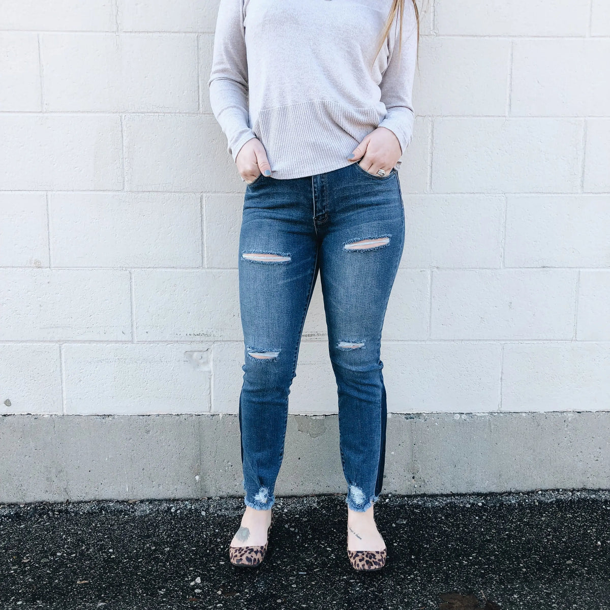 A person, visible from the shoulders down, stands against a white brick wall wearing a light gray long-sleeve top, Hannah Jeggings SIZE 0 with rips, and leopard print flats.