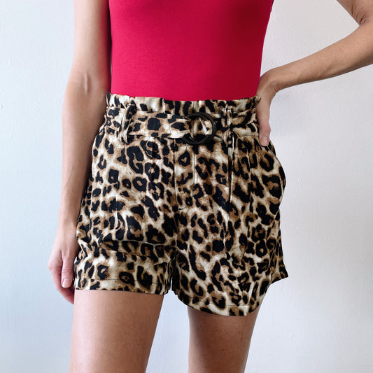 Someone stands against a white background with one hand on their hip, wearing a sleeveless red top and high-waisted Katy Cheetah Shorts featuring a belt buckle.