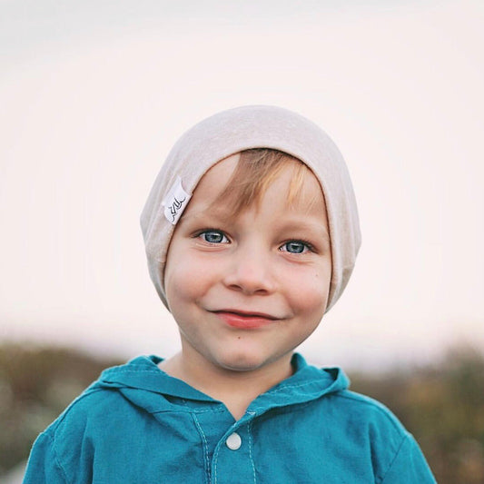 A young child with blue eyes and light brown hair smiles gently, wearing a light tan beanie and a teal shirt, standing outdoors against a softly blurred background.