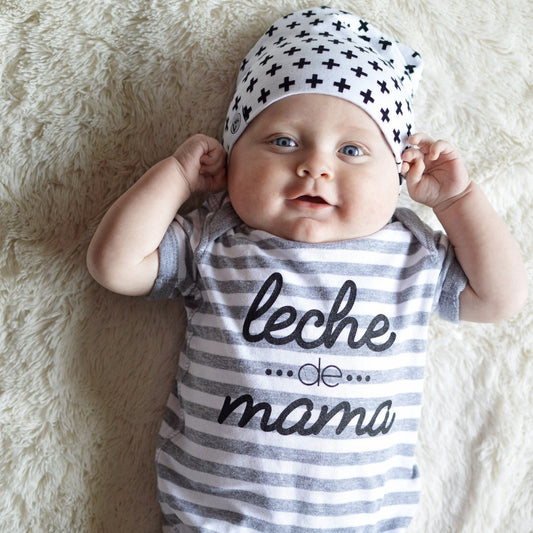 A baby smiles on a soft cream surface, wearing the Leche de Mama Bodysuit—hand-printed in Grand Rapids with gray stripes and leche de mama text—and a white hat with black plus signs, holding the sides of the hat.