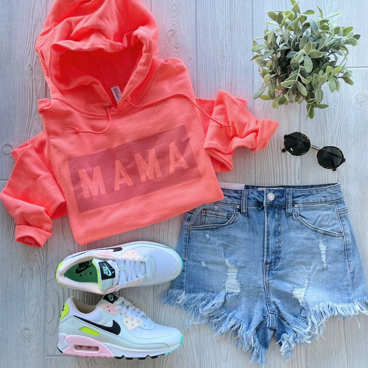 Flat lay of the Mama Box • Neon Tonal Hoodie in a unisex fit labeled MAMA, paired with distressed denim shorts, white Nike sneakers with green accents, black round sunglasses, and a green potted plant on light wooden flooring.