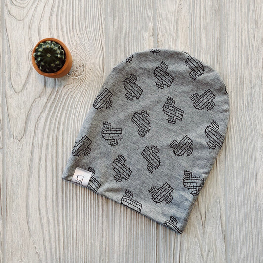 The Cactus Beanie, a handmade gray hat with a black cactus pattern and small white tag, rests on light wood beside a potted cactus—perfect for any River Babe who loves cozy style.
