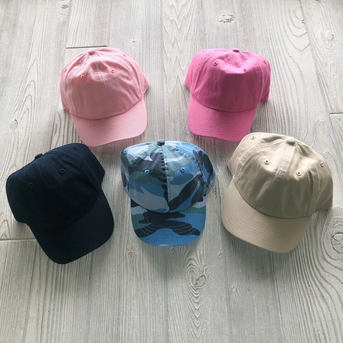 Five caps on a light wooden floor: two pink Kids Baseball Hat • More Colors! at the top, a navy blue cap with an elastic band on the left, a tan cap on the right, and a blue camo-patterned toddler size hat in the center.