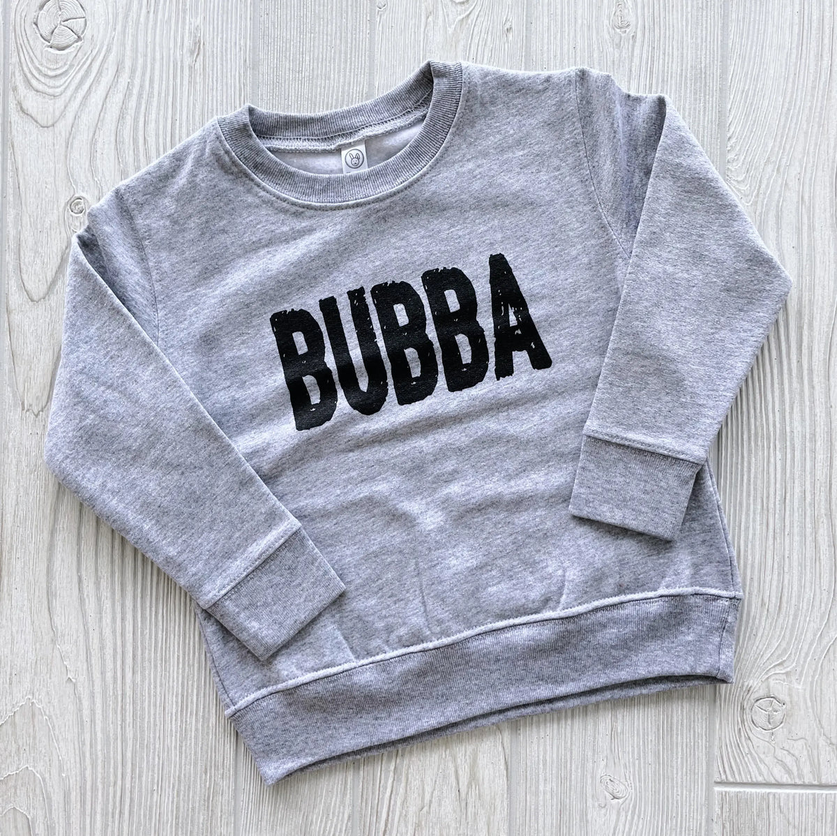 The Bubba Pullover is a comfy, unisex-fit gray sweatshirt with BUBBA in bold black letters across the front, shown laid flat on a light wood surface—perfect for everyday wear.