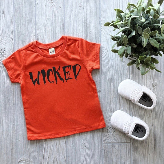 The orange Wicked Tee, featuring WICKED in black spooky letters, is displayed flat on a light wood surface next to white baby shoes and a green potted plant.