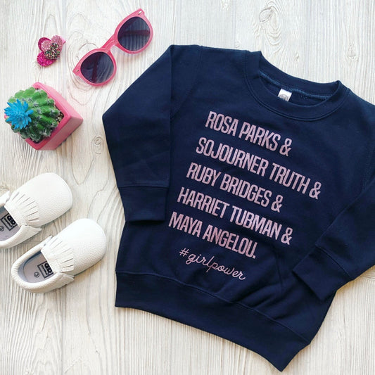 The Girl Power Pullover, a navy blue sweatshirt with white text listing Rosa Parks, Sojourner Truth, Ruby Bridges, Harriet Tubman, and Maya Angelou, honors Black History Month. Styled with white shoes, pink sunglasses, a hairpin & a small cactus.