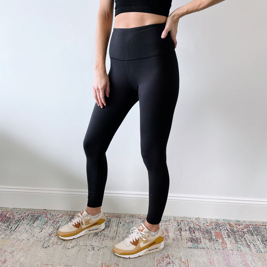 A person in a black crop top, high waist Lennon Leggings, and tan Nike sneakers stands on a patterned rug against a white wall, posing with one hand on their hip.