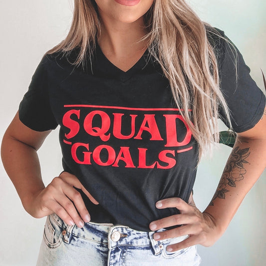 A youth with long blonde hair wears a comfy black Squad Goals • Youth tee in a unisex fit, featuring bold red SQUAD GOALS lettering, paired with light denim jeans. Only the torso is shown, hands placed on hips.
